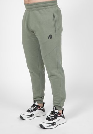 Jogging en olive avec un logo de gorille noir, poches latérales et jambes fuselées. Porté avec des chaussures de sport noires et blanches.