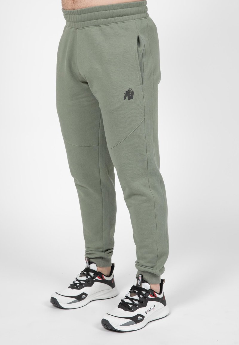 Olijfgroene joggers met een zwart gorilla-logo, zijzakken en taps toelopende benen. Gekozen in combinatie met zwart-witte sportschoenen.