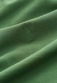 Zöld textília, melynek szövése texturált, finoman hímzett őz logóval díszítve, ami a részletes kézművességre utal.