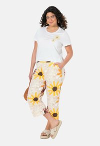 Witte T-shirt met bloemen borduursel, gecombineerd met cropped pants met een multikleuren zonnebloem patroon op een lichte achtergrond. Neutrale sandalen.