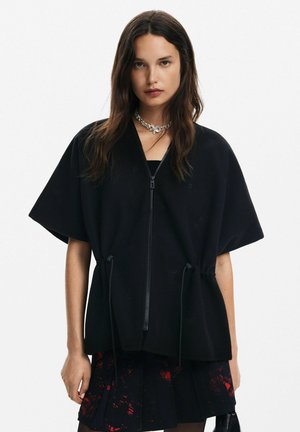 Felpa nera con zip, oversize e maniche corte, dettagli con cordoncino in vita e texture liscia. Indossata sopra una gonna a fantasia.