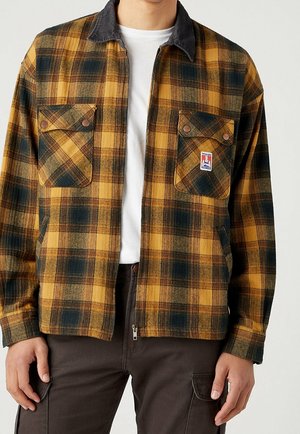 Mann trägt eine senfgelbe und schwarze karierte Jacke mit Vordertaschen, schwarzem Kragen, weißem Hemd darunter und dunkler Hose.