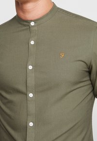 Ciemno oliwkowa koszula na guziki wykonana z bawełny. Ma wysoki kołnierz, białe guziki oraz małe żółte haftowane logo na piersi.