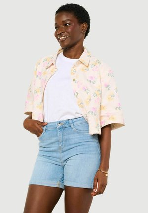 Uśmiechnięta kobieta w lekkiej, kwiecistej koszuli zapinanej na guziki, nosząca ją na białej koszulce i jasnoniebieskich dżinsowych szortach, z jedną ręką w kieszeni i bransoletką na nadgarstku.