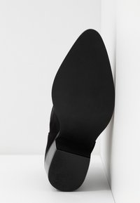 Semelle d'une chaussure élégante noire à bout pointu avec un talon bloc bas, reposant verticalement contre un mur d'angle blanc.