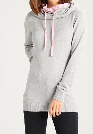 Femme portant un pull gris clair à manches longues avec une capuche rose et des cordons, associée à un pantalon noir, debout avec une main posée sur la cuisse.