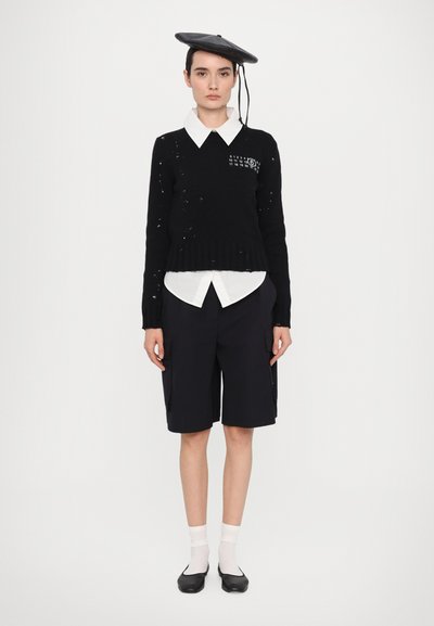 Svart stickad tröja med slitna detaljer, lagd över en vit skjorta med krage, parat med svarta shorts och en svart basker.