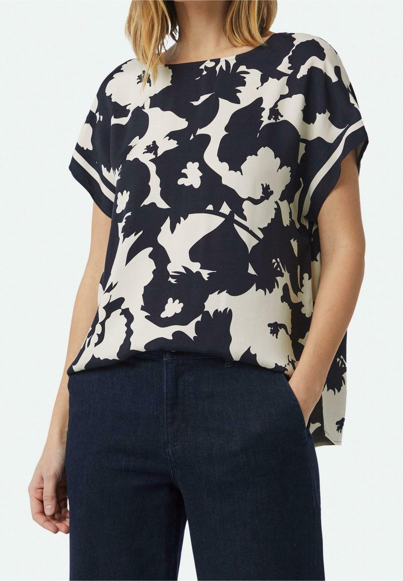 Femme portant un chemisier ample à manches courtes à motif floral noir et blanc, rentré dans un jean bleu foncé, une main dans la poche.