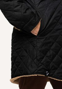 Svart quiltad ytterkläder med en kontrasterande tan fleecefoder vid fållen. Har sidofickor och en logotags längst ner.