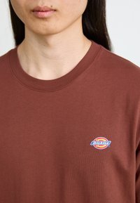 Bruine katoenen crewneck t-shirt met een klein blauw en rood Dickies-logo op de linkerborst. Soelende textuur met een standaard pasvorm.