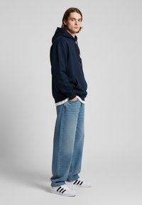 Felpa navy con tasca frontale, abbinata a jeans larghi azzurri e sneakers bianche con strisce nere. Design semplice e casual.
