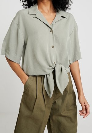 Jasnozielona koszula z krótkim rękawem, z kołnierzykiem, zapinana na guziki z przodu, zawiązana w węzeł na talii, zestawiona z oliwkowymi spodniami o wysokim stanie.