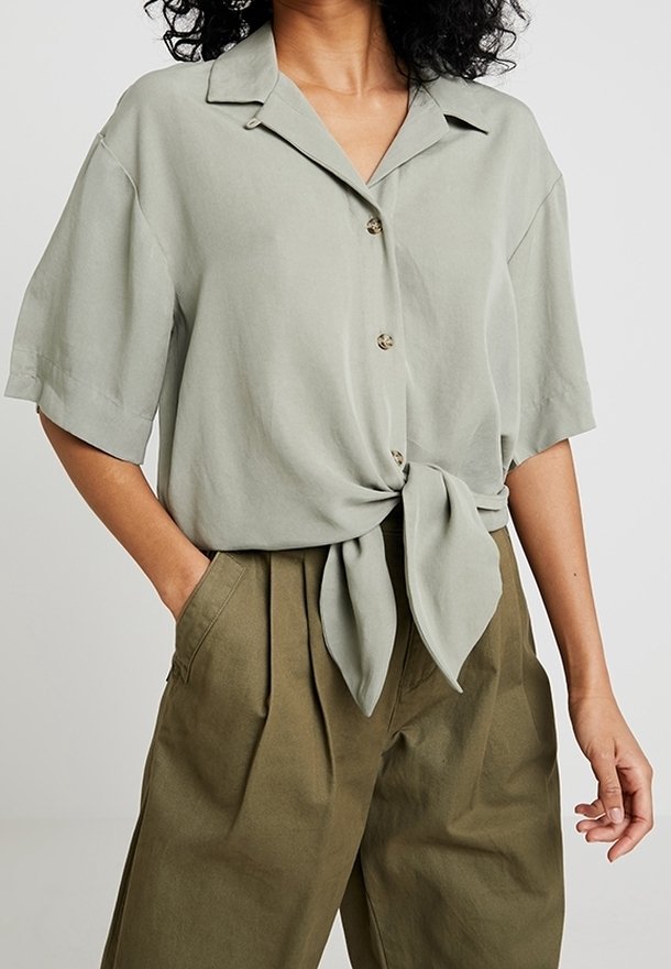 Chemisier vert clair à manches courtes avec col, boutonnage sur le devant et nœud noué à la taille, associé à un pantalon taille haute de couleur olive.