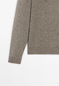 Manche et côté d'un pull en maille brun mouchetée, bord-côtes aux poignets et à l'ourlet visibles, posés à plat sur un fond blanc.