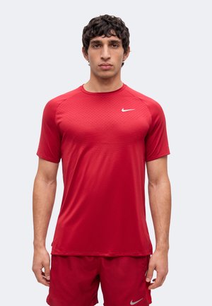 Mladý muž s tmavými kučeravými vlasmi, ktorý má na sebe červené športové tričko Nike s krátkym rukávom a zodpovedajúce červené šortky, stojí čelom vpred.