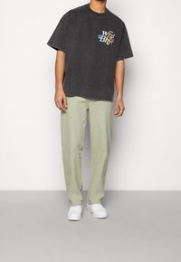 Grå oversized t-shirt med flerfärgad "Worst Behavior" logotyp, tillsammans med ljusgröna lösa byxor och vita sneakers.