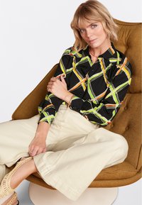 Zwarte shirt met kleurrijk geometrisch patroon in groen, geel en oranje. Model draagt een crème wijde broek en gevlochten sandalen, zittend op een stoel.