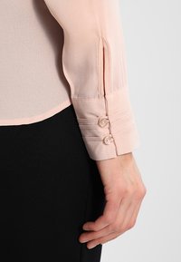 Light pink blouse with a sleek, sheer fabric. The cuff features two buttons and striped accents along the sleeve. Paired with a black garment.