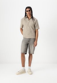 Camicia a maniche corte a quadri in toni beige, abbinata a pantaloni corti in denim grigi e sneaker color crema. Il modello indossa occhiali da sole rotondi e ha i capelli lunghi.