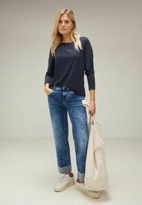 Marineblaues Langarmoberteil mit geprägtem Text, kombiniert mit hellblauen, taillierten Jeans mit Säumen und weißen Sneakers. Hält eine helle Tragetasche.