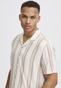 Camisa de botones de manga corta hecha de tela ligera, con rayas verticales en tonos crema y marrón, con un cuello relajado y una textura sutil.