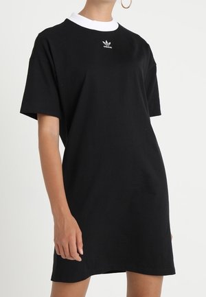 Kvinde iført en kort sort Adidas-kjole med hvid krave og logo, stående foran en ensfarvet lys baggrund.