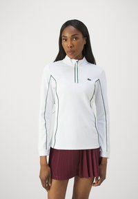 Camisa blanca de manga larga con detalles verdes y cuello con media cremallera, combinada con pantalones cortos de pliegues en color burdeos. Fabricada con una tela suave.