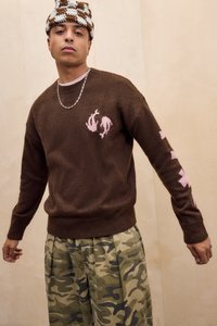 Brauner Fleece-Pullover mit pinkem Herzdesign und Akzenten an den Ärmeln, kombiniert mit grünen Camouflage-Hosen mit lockerem Schnitt und Falten.