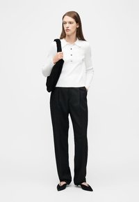 Femme portant une chemise blanche à manches longues avec col, un pantalon noir à jambes larges, des chaussures noires à bouts pointus, et un sac à bandoulière noir texturé.