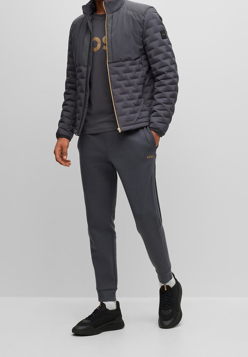 Homme portant une veste matelassée gris foncé, un pantalon de survêtement assorti avec le logo BOSS, des baskets noires et une chemise sombre avec un motif doré, debout devant un fond gris.