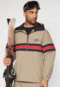 Μπεζ σκι Jacket με μαύρη και κόκκινη ρίγα, με φερμουάρ μπροστά και λογότυπο. Άτομο κρατά σκι, φορώντας μαύρα γυαλιά σκι.