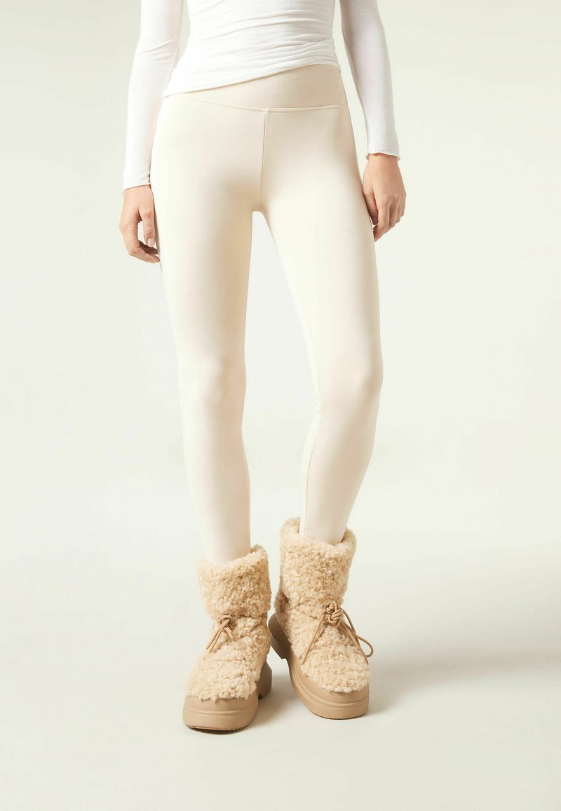 Leggings beige con un diseño sin costuras combinados con botines de pelaje marrón claro que tienen una suela gruesa y cordones atados. Se nota una textura suave.