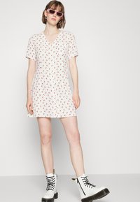 Vestido branco com padrão floral vermelho, mangas curtas, decote em V e um corte solto. Usado com botas brancas de atacadores e óculos de sol cor-de-rosa.