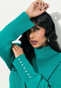 Türkiser Pullover mit geripptem Rollkragen und Bündchen. Das Bündchen hat sechs perlenartige Knöpfe. Weicher Strickstoff, oversized Schnitt.