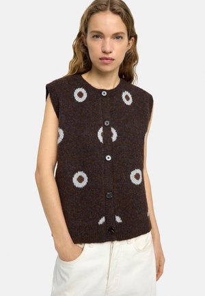 Kobieta ubrana w bezrękawnik wykonany z ciemnobrązowego dzianego materiału z szarymi okrągłymi wzorami i czarnymi guzikami, w połączeniu z jasnymi spodniami.