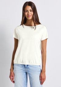Top blanco de manga corta con escote redondeado, detalles texturizados en el hombro y dobladillo gather. Combinado con jeans azul claro.