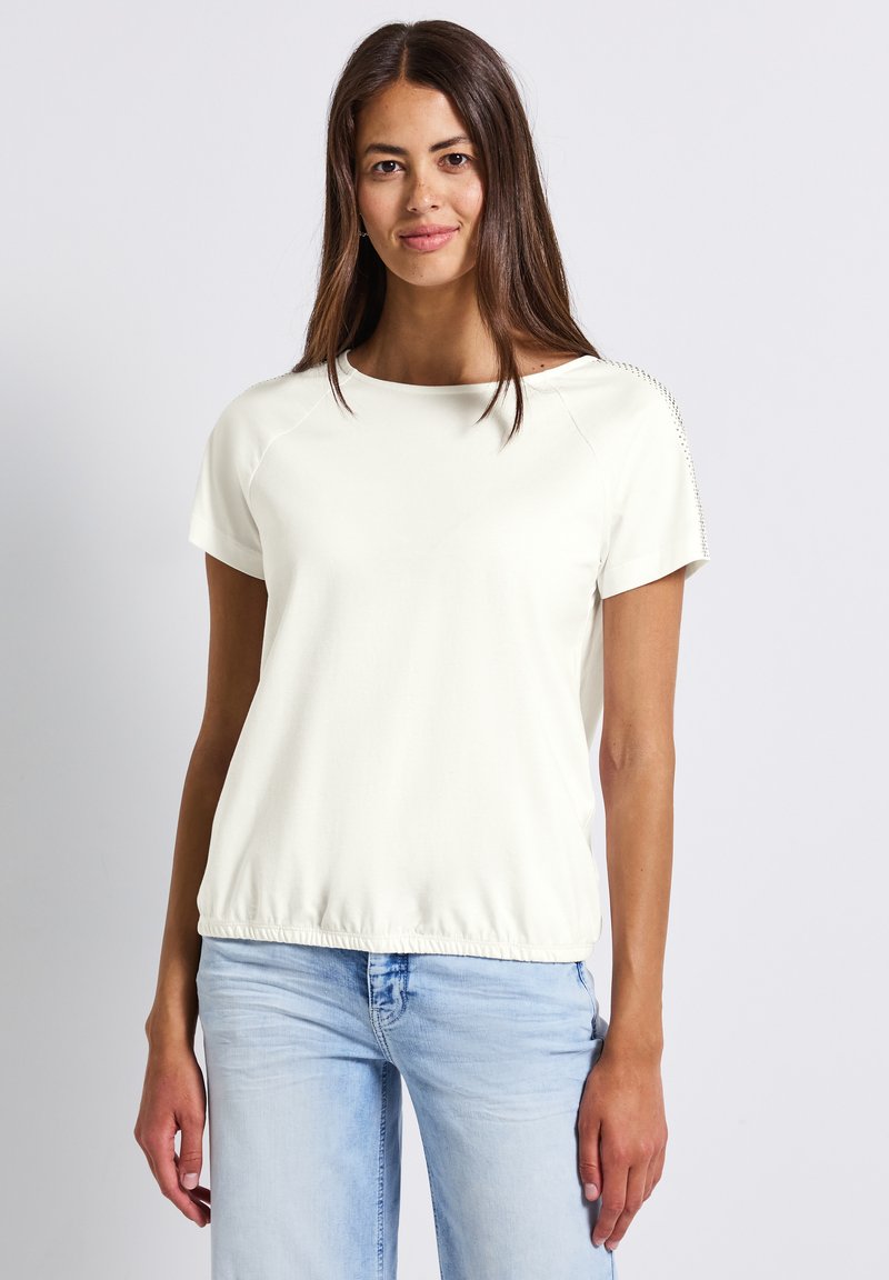 Top blanco de manga corta con escote redondeado, detalles texturizados en el hombro y dobladillo gather. Combinado con jeans azul claro.
