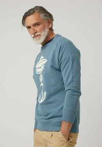 Hombre de mediana edad con cabello y barba canosos, vistiendo una sudadera azul con un gráfico de ancla, pantalones beiges y manos en los bolsillos, frente a un fondo simple.
