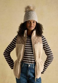 Tan quilted vest with cream sherpa panels, worn over a brown and white striped long-sleeve shirt. Finished with blue jeans and a knitted hat.