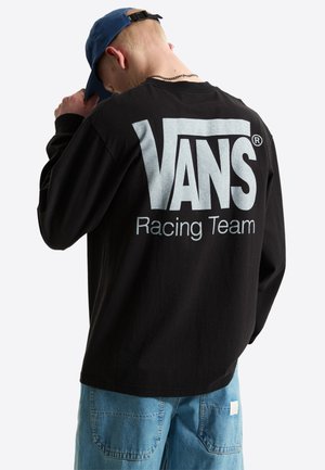 Osoba w czarnej, długiej koszulce Vans Racing Team i niebieskiej czapce, zwrócona tyłem, widoczne jasnoniebieskie dżinsy.