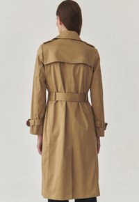 Khaki trench coat made of smooth fabric with a belted waist, epaulettes, and back vent, featuring long sleeves and a classic silhouette.