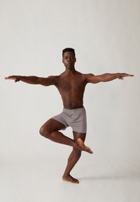 Grijze katoenen boxershorts op een persoon in een evenwichtige yogahouding, met uitgestrekte armen, voor een neutrale achtergrond en blote voeten in beeld.