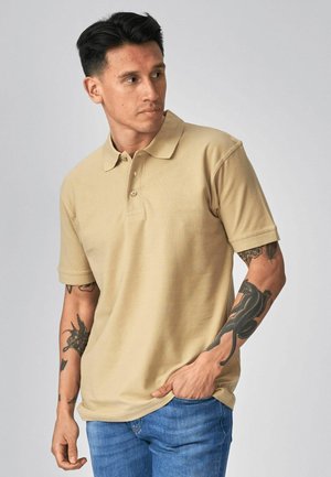 Fekete hajú férfi bézs színű pólóingben és kék farmerral, mindkét karján tetoválásokkal, oldalra néz, az egyik kezét a zsebében tartva.