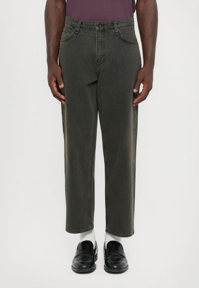 Homme portant un jean court gris charbon, des chaussettes blanches, des mocassins en cuir noirs et une chemise violette rentrée, se tenant devant un fond uni.