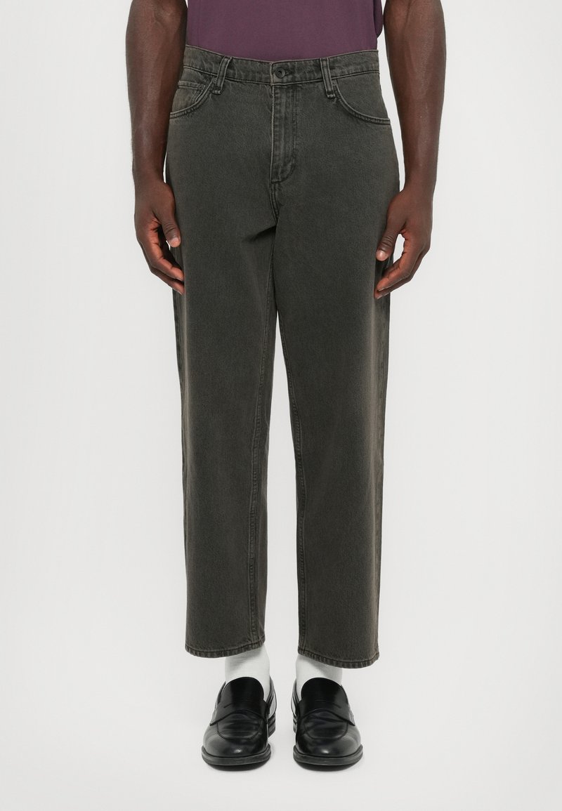 Man med kolgrå avklippta jeans, vita strumpor, svarta läderskor och en lila skjorta instoppar, som står framför en neutral bakgrund.