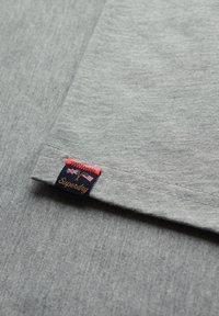 Tela de camiseta de algodón gris con un borde doblado, que presenta una etiqueta "Superdry" roja y azul cosida en el dobladillo. Textura suave, diseño casual.