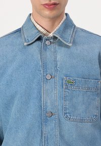 Mann trägt hellblaue Jeansjacke mit Knopfverschluss und Brusttasche mit kleinem grünem Krokodil-Logo-Aufnäher.