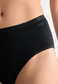 Close-up van de taille en heup van een persoon die zwarte hoge taille onderbroek draagt met een klein "Sloggi" logo op de tailleband.