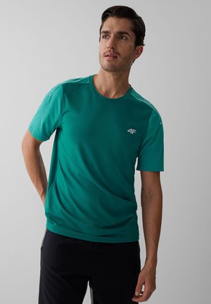 Homme aux cheveux foncés portant un maillot de sport à manches courtes bleu sarcelle et un pantalon noir, regardant légèrement de côté sur un fond gris uni.