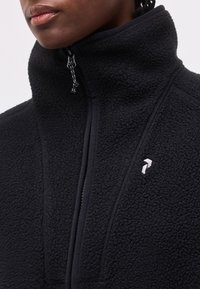 Κοντινό πλάνο προσώπου που φοράει μαύρο φλις μπουφάν με ψηλό γιακά και μικρό λευκό λογότυπο στο στήθος, μερικώς φερμουάρ ανοιχτό.